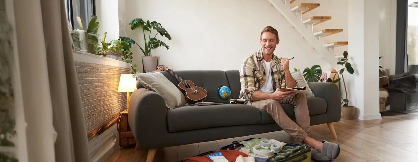 Счастливый мужчина с ручкой и блокнотом проверяет список вещей и упаковывает чемодан для поездки, собираясь в отпуск и готовясь к летним каникулам, сидя в гостиной.